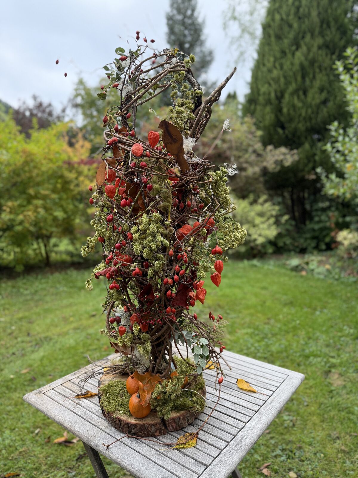 Hohes Herbstgesteck mit Lampionblumen, Hagebutten, Hopfen, Rostvogel und kleinen Zierk&uuml;rbissen auf Baumscheibe